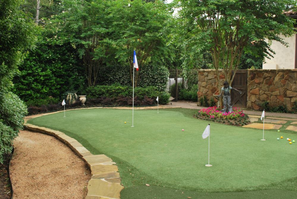 Backyard putting green made with artificial grass, featuring multiple golf holes and flags, surrounded by landscaped trees, stone edging, and a garden walkway.